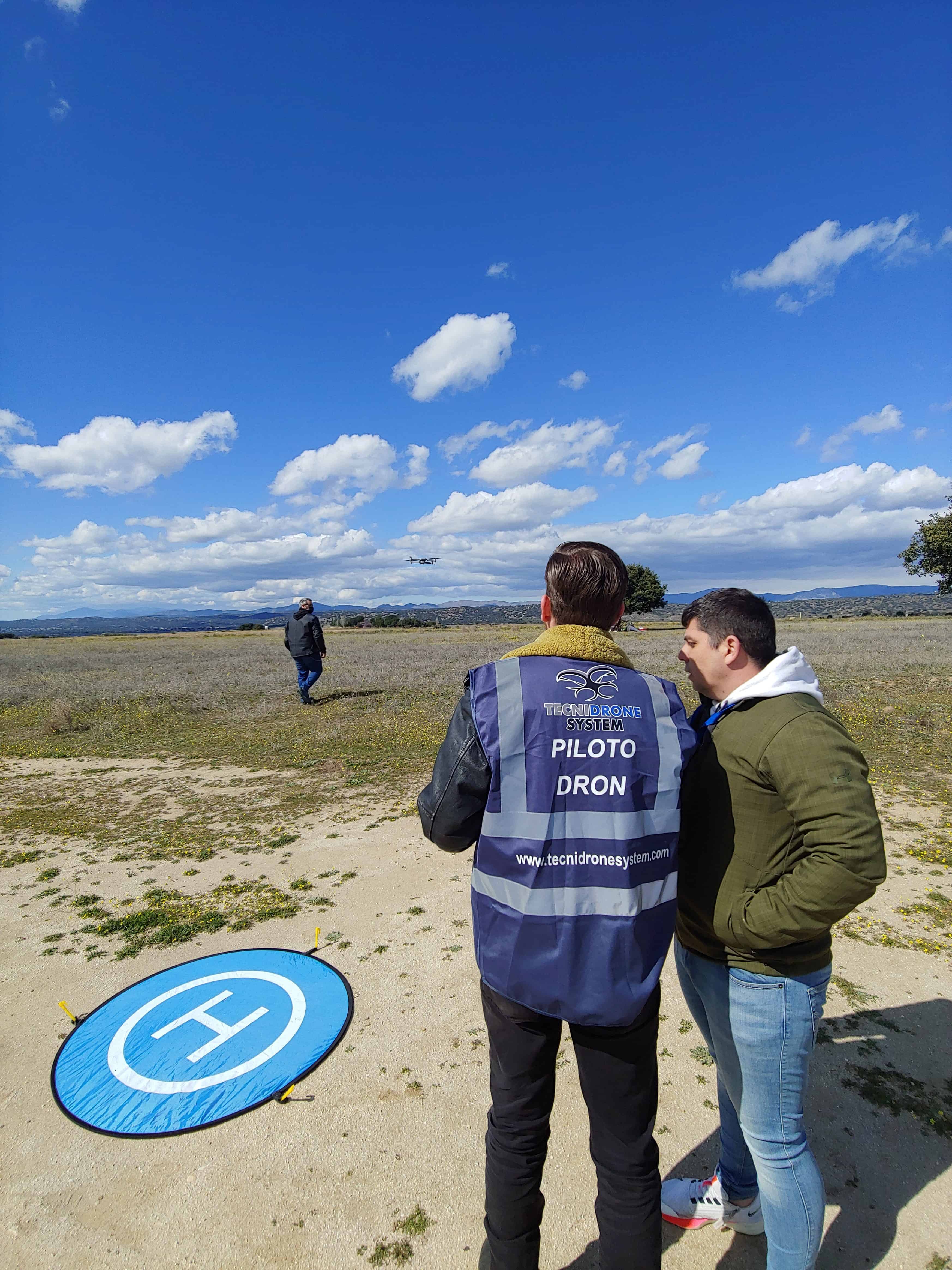 Un piloto de TecniDrone con chaleco de piloto de dron durante una práctica en campo, con el dron volando al fondo