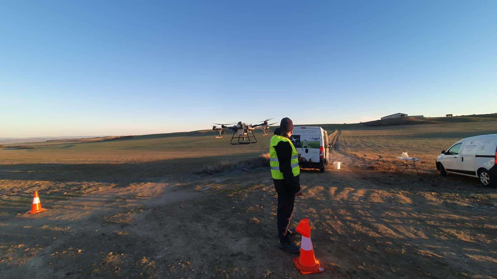 Un piloto con chaleco reflectante vuela un dron agrícola en campo abierto, junto a dos furgonetas